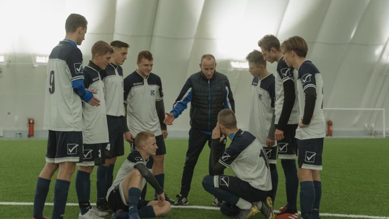 A soccer coach gives tactical advice to a youth team indoors on artificial turf.