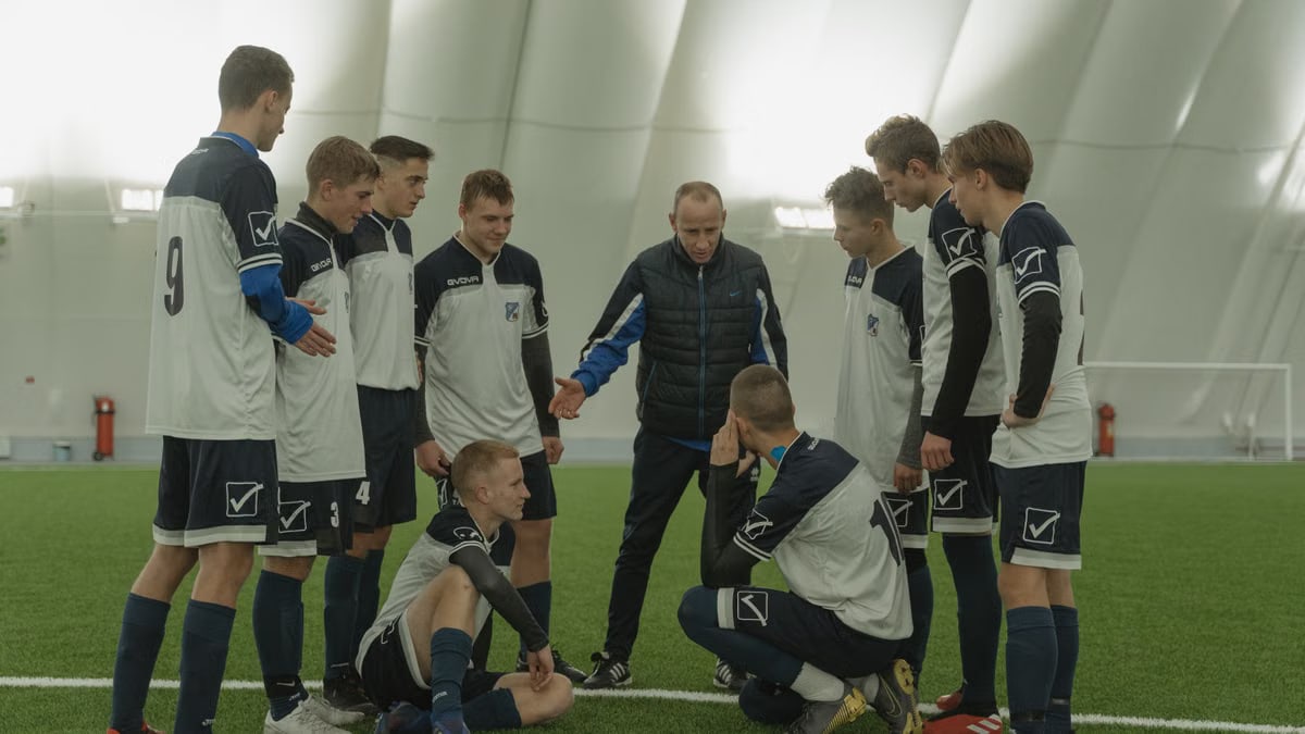 A soccer coach gives tactical advice to a youth team indoors on artificial turf.
